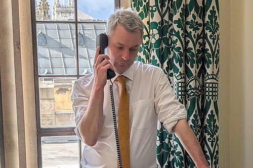 Will Forster speaking on the phone in his Westminster office, he is wearing a white shirt with the sleeves rolled up and a yellow tie.