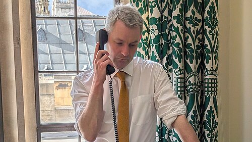 Will Forster speaking on the phone in his Westminster office, he is wearing a white shirt with the sleeves rolled up and a yellow tie.