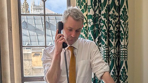 Will Forster speaking on the phone in his Westminster office, he is wearing a white shirt with the sleeves rolled up and a yellow tie.