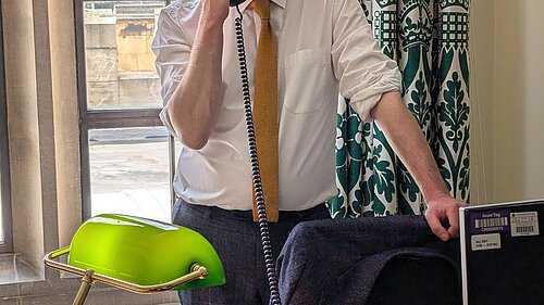 Will Forster speaking on the phone in his Westminster office, he is wearing a white shirt with the sleeves rolled up and a yellow tie.